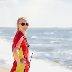 Livräddare i rött och gult står på stranden, redo för sol och bad. Vågorna skvalpar vid hennes fötter.