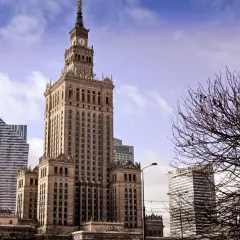 Kulturpalatset Warszawa reser sig stolt mot en molnig himmel, omgiven av moderna skyskrapor och kala trädgrenar.