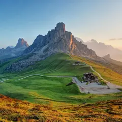 Vacker solnedgång över Dolomiterna. En slingrande väg leder upp till ett hus på en grön kulle, perfekt för en vandring.
