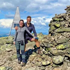 Två glada vandrare vid en stenröse på en bergstopp i Sverige. En modern, pyramidformad byggnad med solpaneler syns i bakgrunden. Perfekt för att utforska Sveriges vandringsleder.