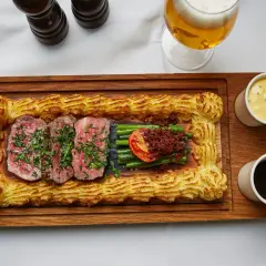 Sliced steak with green beans, tomato, and mashed potato border on a wooden board, with beer and sauces.