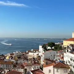 Vy över Lissabon gamla stan med röda tak, en stor kryssningsbåt och en mindre båt på det blå vattnet under en klarblå himmel.