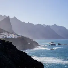 En vit by klättrar på en grön sluttning vid havet, med dramatiska berg i bakgrunden. En dröm om flygtid Teneriffa.