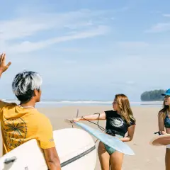 Tre vänner med surfbrädor på stranden i Thailand. En man med vitfärgat hår vinkar, medan två kvinnor förbereder sig för att surfa.