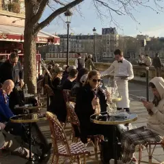 En solig dag på en restaurang i Paris. Människor sitter vid borden utomhus, och servitörer bär brickor med mat och dryck.