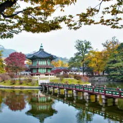 En vacker höstbild från Seoul, Sydkorea. Ett traditionellt koreanskt pagodliknande hus står på en ö i en damm, med en bro som leder dit. Träden runt omkring har höstfärger.