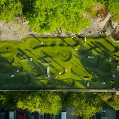 Folk spelar minigolf i Nacka Strand. En grön bana med hinder och skuggor från träden.