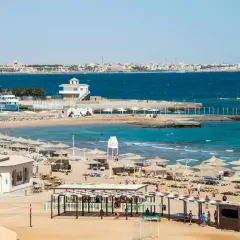 Solig strand i Hurghada, Egypten, med parasoller, solstolar och turkost vatten. En båt ligger vid en pir.