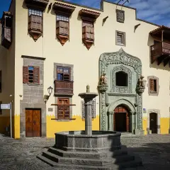 En vacker fontän framför en gul byggnad med träbalkonger och fönsterluckor i Las Palmas gamla stan.