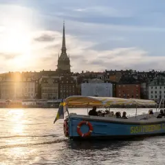 A blue sightseeing boat with a yellow striped canopy sails on water in Stockholm, with historic buildings and a church spire in the background at sunset.