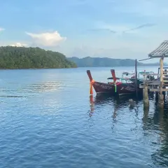 Två longtailbåtar vid en träbrygga på Koh Jum. En grönskande ö syns i bakgrunden under en molnig himmel.