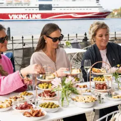 Kvinnor njuter av mat och dryck vid ett bord på en sq restaurang med Viking Line-färja i bakgrunden.