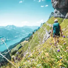 Vandra i Schweiz längs en bergssluttning med en turkos sjö nedanför. Blommor i förgrunden, en vandrare med ryggsäck på väg uppåt.