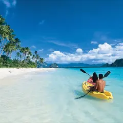 Två personer paddlar kajak i det turkosa vattnet vid en vit sandstrand med palmer på Palawan, Filippinerna.