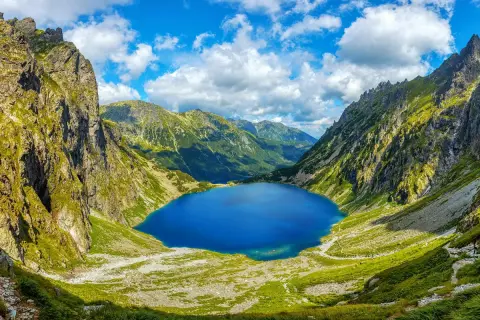 Blågrönt Morskie Oko, en sjö omgiven av gröna bergssluttningar och dramatiska klippor under en molnig himmel.