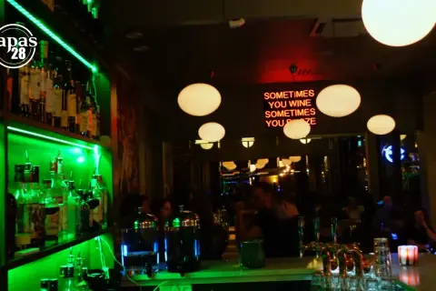 En livlig bar med grönt ljus som lyser upp hyllor med flaskor. En neonskylt med texten "SOMETIMES YOU WINE SOMETIMES YOU POZE" hänger ovanför en bar där gäster sitter. Tapas 28.