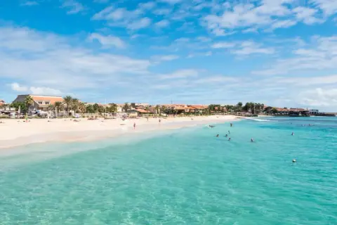 En vacker strand med turkost vatten och palmer. Människor badar och solar. En vy som påminner om en semester vid kap verde flygplats.