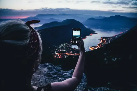 En person tar en bild med mobilen av en stad vid havet i Europa, med berg i bakgrunden.