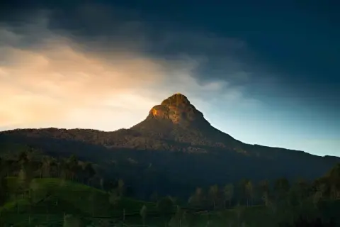 Adam's Peak reser sig majestätiskt mot en dramatisk himmel. Bergssluttningarna är täckta av frodig grönska och teodlingar.