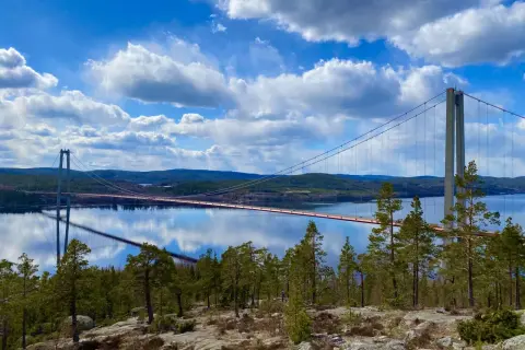 A suspension bridge spans a calm body of water under a bright blue sky with fluffy white clouds.