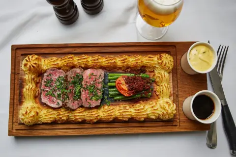 Sliced steak with green beans, tomato, and mashed potato border on a wooden board, with beer and sauces.