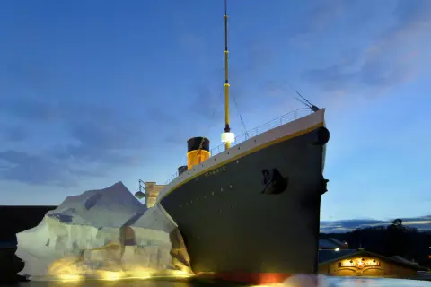 A replica of the Titanic ship is shown at dusk, with an iceberg and the White Star Line building.