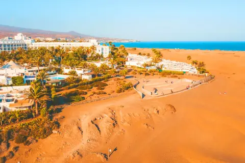 Vacker vy över Dunas de Maspalomas med vita hotell, palmer och gyllene sanddyner mot det blå havet.