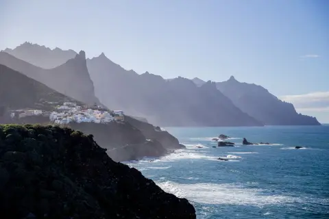 En vit by klättrar på en grön sluttning vid havet, med dramatiska berg i bakgrunden. En dröm om flygtid Teneriffa.