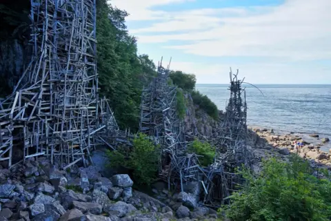 Lars Vilks Nimis, en skulptur av trä som reser sig mot himlen vid kusten.