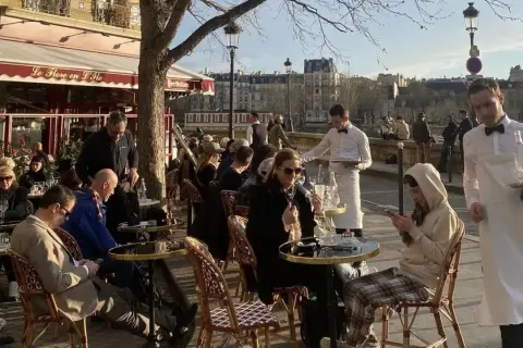 En solig dag på en restaurang i Paris. Människor sitter vid borden utomhus, och servitörer bär brickor med mat och dryck.