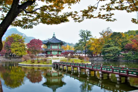 En vacker höstbild från Seoul, Sydkorea. Ett traditionellt koreanskt pagodliknande hus står på en ö i en damm, med en bro som leder dit. Träden runt omkring har höstfärger.
