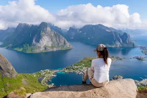 En person sitter på en klippa och blickar ut över en dramatisk kustlinje med berg och en by. En fantastisk vy för lofoten vandring.