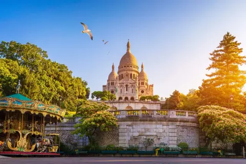Sacre Coeur reser sig majestätiskt mot en klarblå himmel, med en karusell i förgrunden och en mås som svävar ovanför.