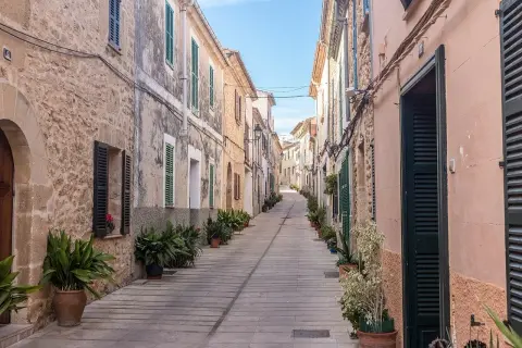 En smal, stenlagd gata i Alcudia old town kantas av gamla hus med gröna fönsterluckor och krukväxter.