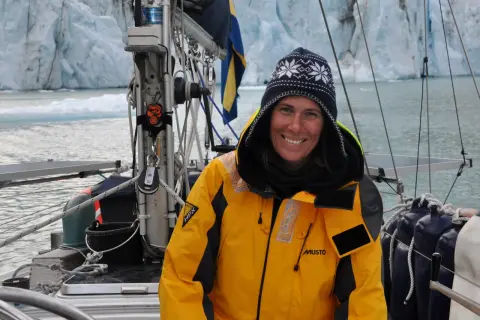 Glad kvinna i gul jacka på segelbåt framför glaciär. Drömmen att segla jorden runt är nära.