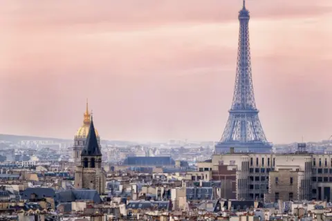 Eiffeltornet höjd över Paris takåsar i rosa skymningsljus. En gyllene kupol syns i bakgrunden.