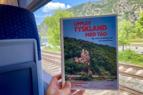 En hand håller en bok om att uppleva Tyskland med tåg, med en slående bild av ett slott vid en flod. Utsikten från tågfönstret visar räls och gröna kullar.