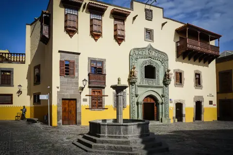 En vacker fontän framför en gul byggnad med träbalkonger och fönsterluckor i Las Palmas gamla stan.