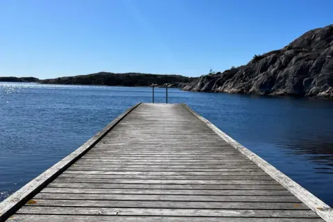 Träbrygga leder ut mot det glittrande vattnet vid Grebbestad strand. Klippor och öar i bakgrunden under en klarblå himmel.
