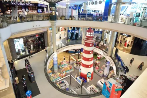 Inne i Terminal 21 Bangkok, ett shoppingcenter med flera våningar, syns en stor rödvit fyr i mitten. Folk shoppar och rör sig mellan butiker som Victoria's Secret.