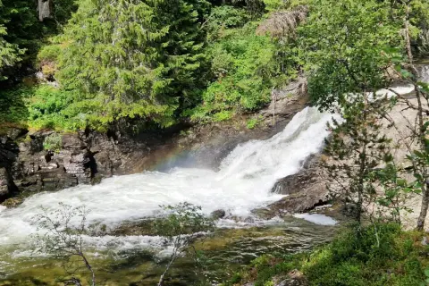 Vattenfall i Idre, med en regnbåge som skimrar i dimman från det forsande vattnet. Omges av grönskande träd och klippor.