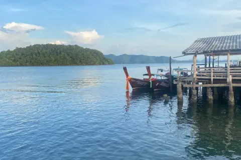 Två longtailbåtar vid en träbrygga på Koh Jum. En grönskande ö syns i bakgrunden under en molnig himmel.