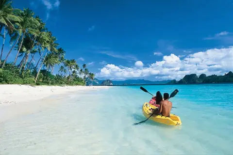 Två personer paddlar kajak i det turkosa vattnet vid en vit sandstrand med palmer på Palawan, Filippinerna.