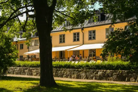 En gul byggnad med vita markiser och uteservering. En lång rad med träd skymmer delvis vyn.
