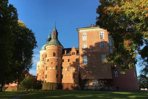 A red brick medieval castle with green domed towers under a clear blue sky, framed by autumn trees.