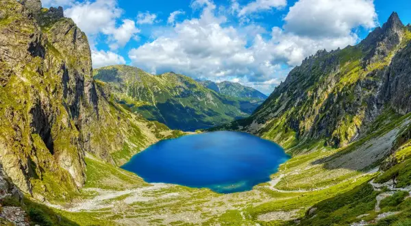 Blågrönt Morskie Oko, en sjö omgiven av gröna bergssluttningar och dramatiska klippor under en molnig himmel.