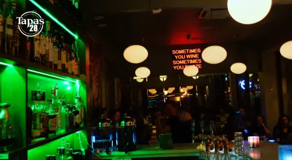 En livlig bar med grönt ljus som lyser upp hyllor med flaskor. En neonskylt med texten "SOMETIMES YOU WINE SOMETIMES YOU POZE" hänger ovanför en bar där gäster sitter. Tapas 28.