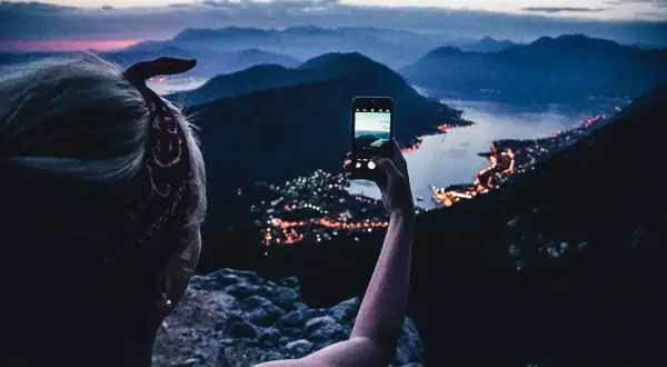 En person tar en bild med mobilen av en stad vid havet i Europa, med berg i bakgrunden.
