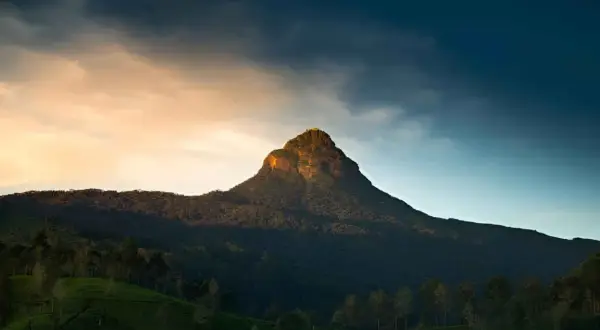 Adam's Peak reser sig majestätiskt mot en dramatisk himmel. Bergssluttningarna är täckta av frodig grönska och teodlingar.