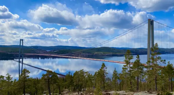 A suspension bridge spans a calm body of water under a bright blue sky with fluffy white clouds.